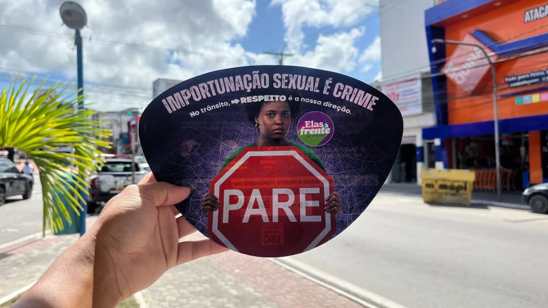 Na imagem uma mão segura um folheto triangular com bordas arredondadas aonde há uma mulher negra segurando a placa escrito "PARE" e acima o titulo "Importunação Sexual é Crime"