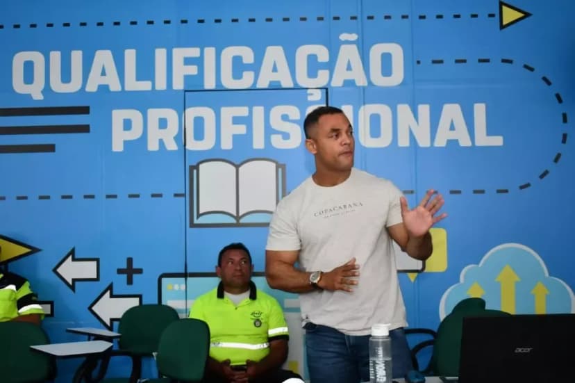 Na imagem, um professor pardo com camisa branca e calça jeans, explicando na aula de qualificação profissional os agentes de trânsito. 