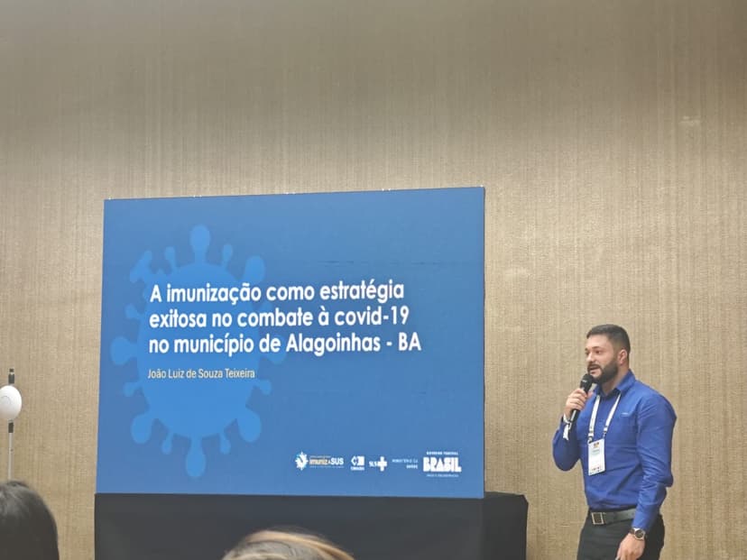 Imagem referente a oficina Nacional do ImunizaSUS. Na imagem há um slide em azul e um homem de cor branca, camisa azul segurando um microfone